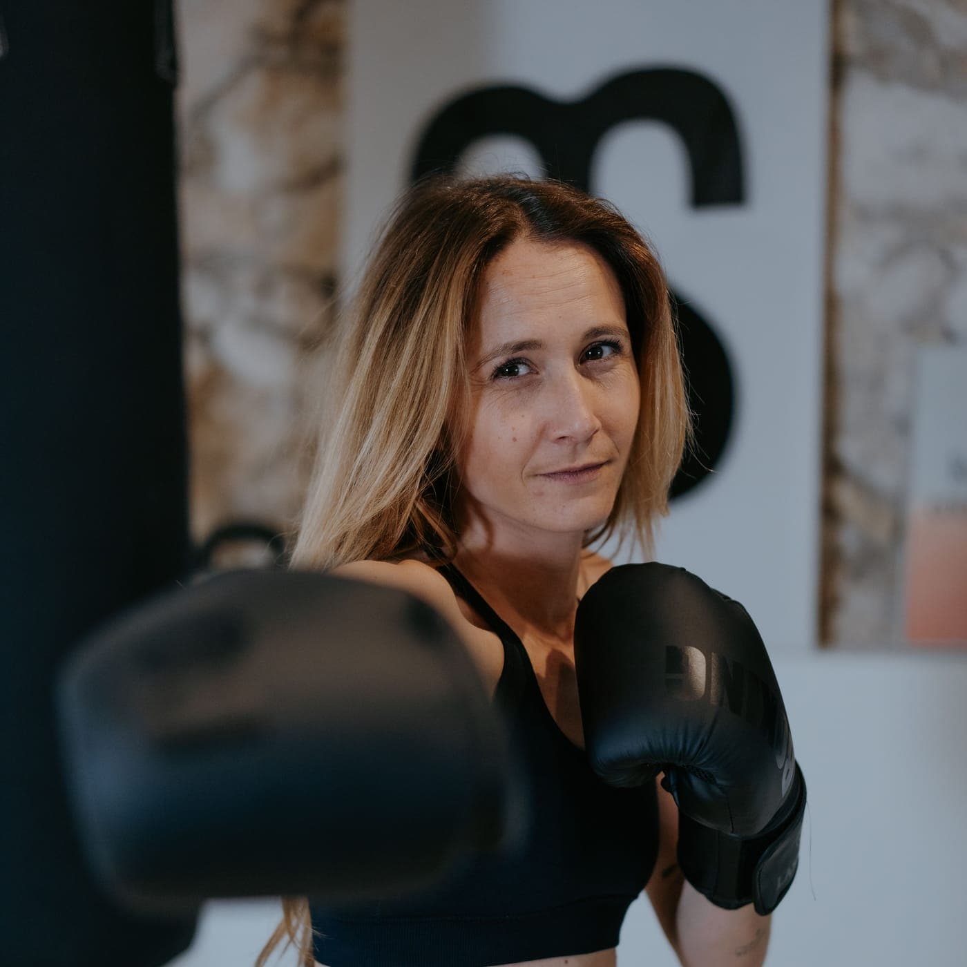 Coach du Club Sisko en garde, gants de boxe noirs, devant le sac de frappe et l'affiche au monogramme du club